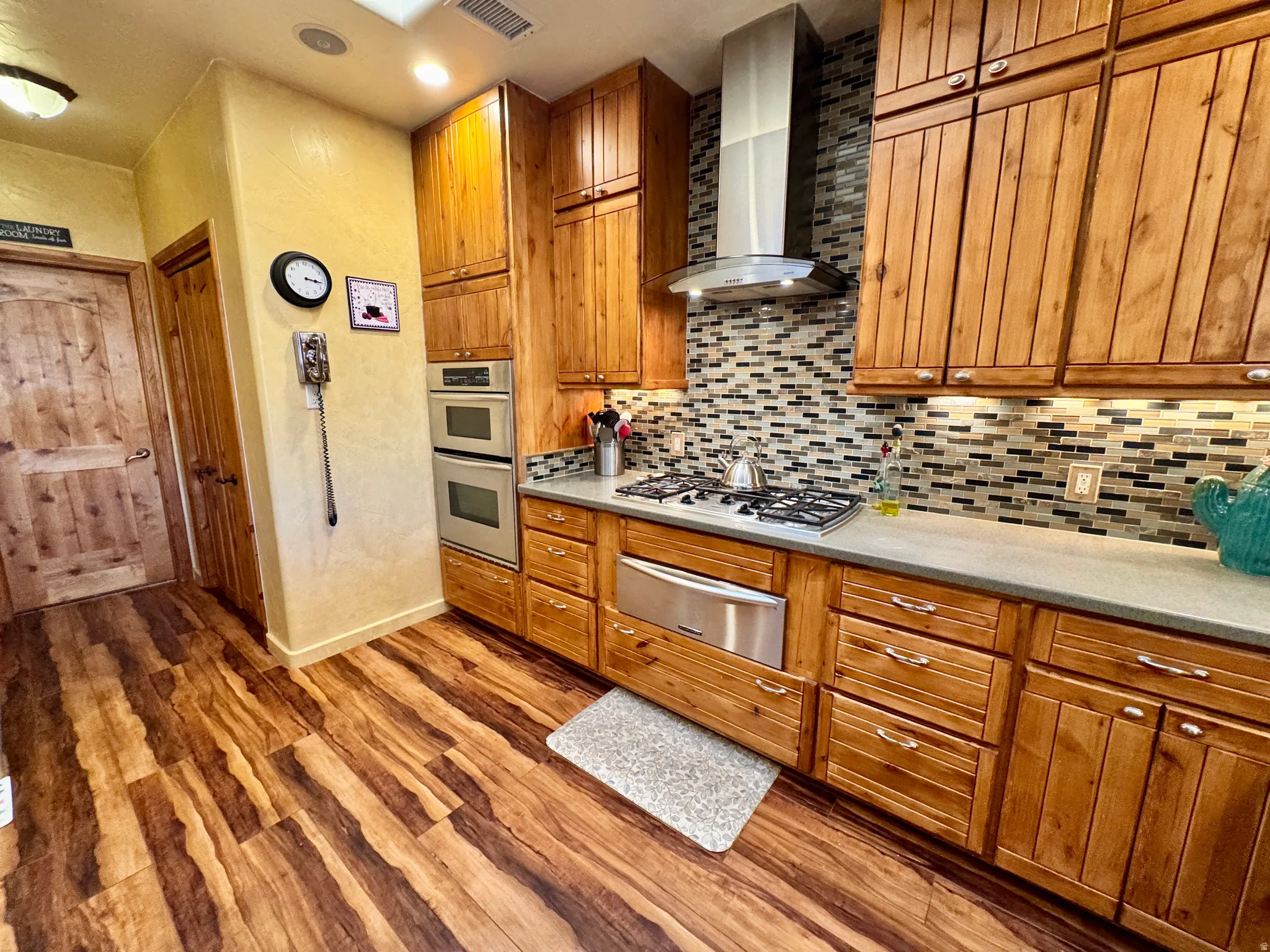 Kitchen featuring brown cabinetry, wall chimney exhaust hood, a warming drawer, dark wood-style flooring, and recessed lighting