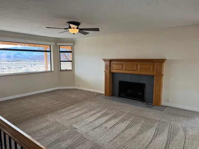 Unfurnished living room with carpet, a tiled fireplace, a textured ceiling, crown molding, and a ceiling fan