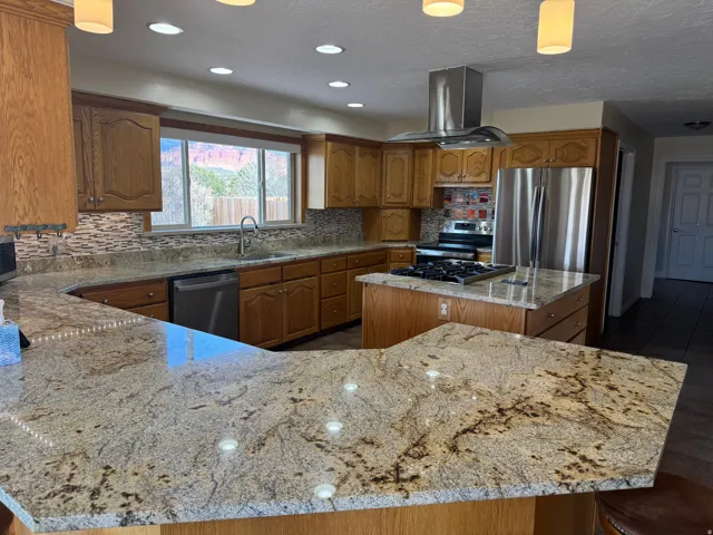 Kitchen with a kitchen island, brown cabinetry, light stone counters, hanging light fixtures, and appliances with stainless steel finishes