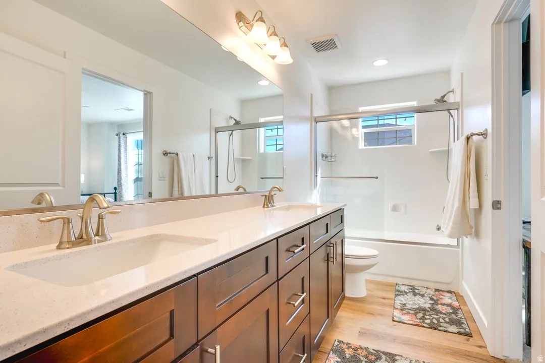 Full bath featuring double vanity, light wood-style flooring, enclosed tub / shower combo, and recessed lighting
