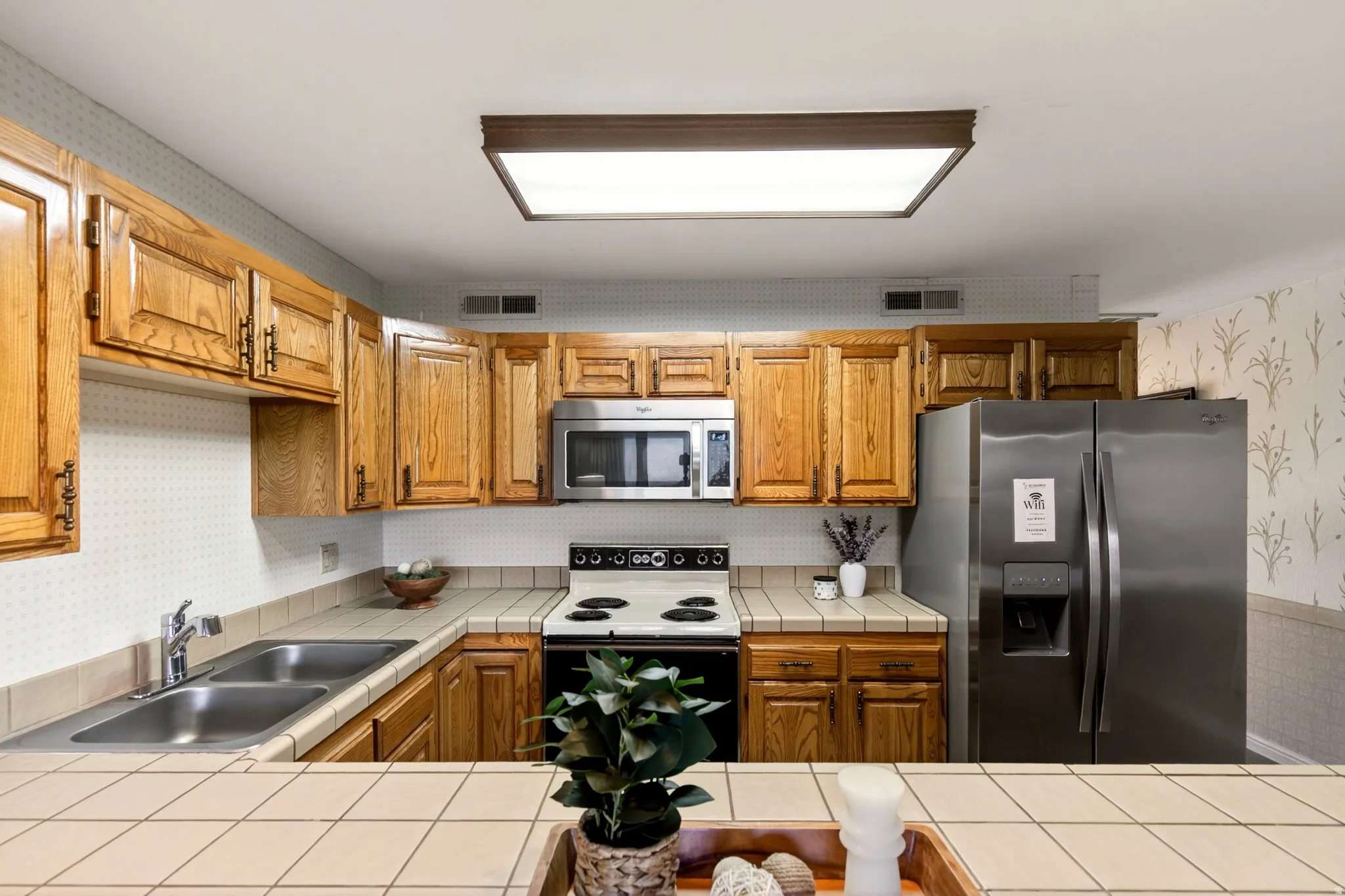 Kitchen featuring wallpapered walls, appliances with stainless steel finishes, brown cabinetry, and tile countertops