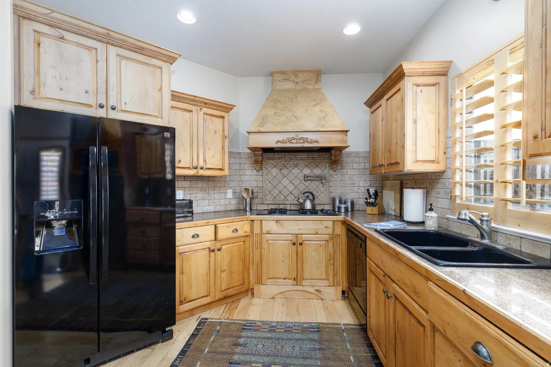 Kitchen with black appliances, custom range hood, tasteful backsplash, light wood-style flooring, and light brown cabinets