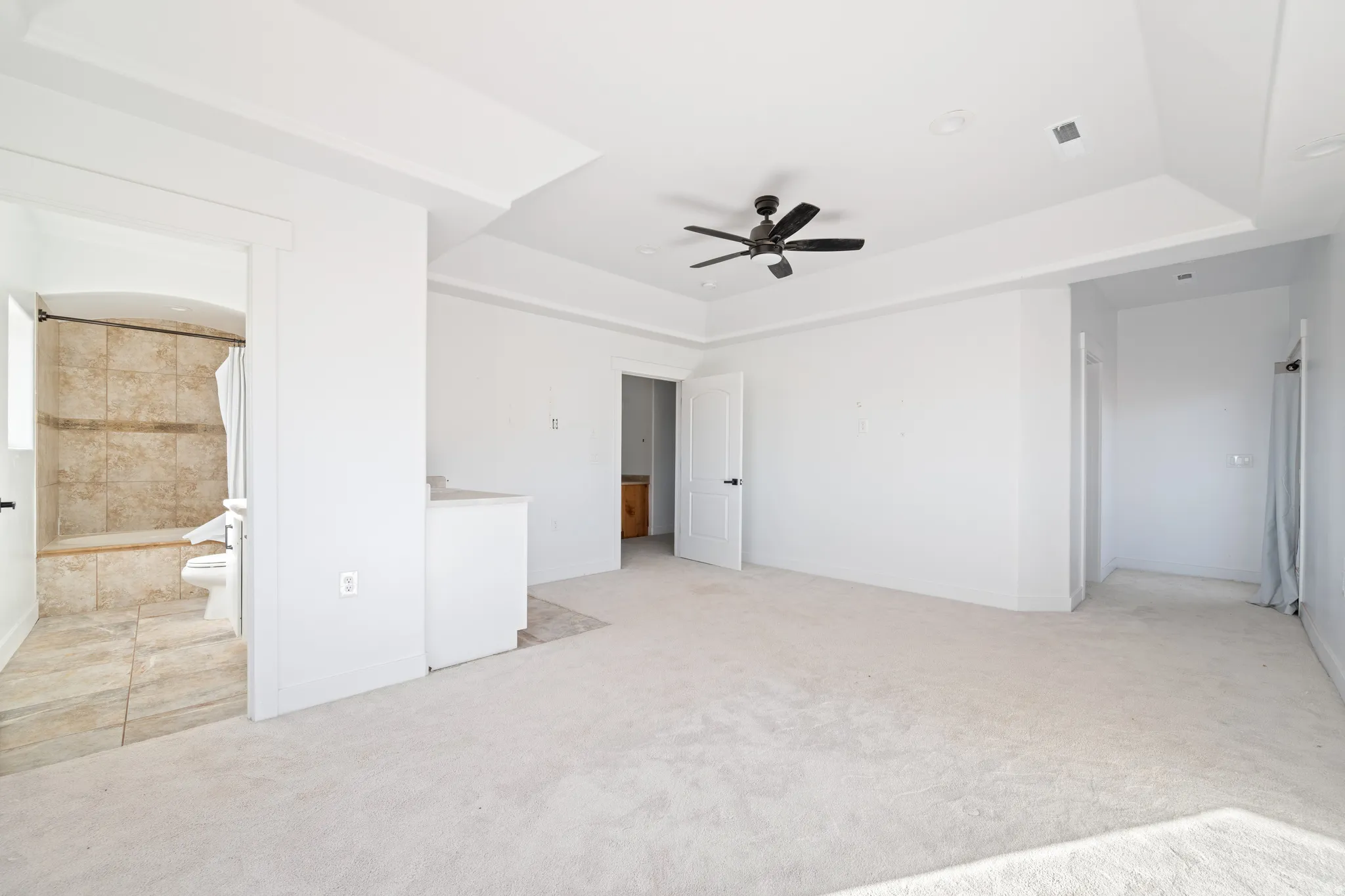 Unfurnished bedroom featuring light colored carpet, ceiling fan, ensuite bath, and a tray ceiling