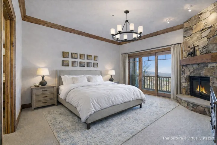 Bedroom featuring a stone-clad fireplace with wood mantle, private balcony access through glass doors, exposed wooden ceiling beams, and a black metal chandelier