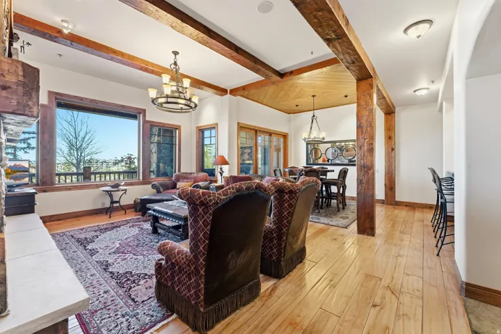 Spacious living area featuring exposed wood beams, hardwood flooring, a large picture window, a stone fireplace, and a decorative chandelier