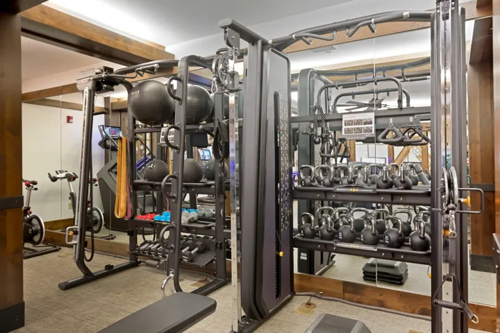 Dedicated fitness space featuring mirrored walls, wood-finish structural beams, carpet flooring, a multi-station exercise machine, and kettlebell storage