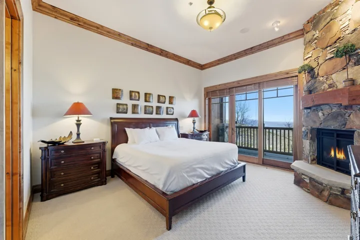 Spacious bedroom featuring wood-trimmed vaulted ceilings, a stone-faced fireplace with a wood mantel, and expansive glass doors opening to a balcony