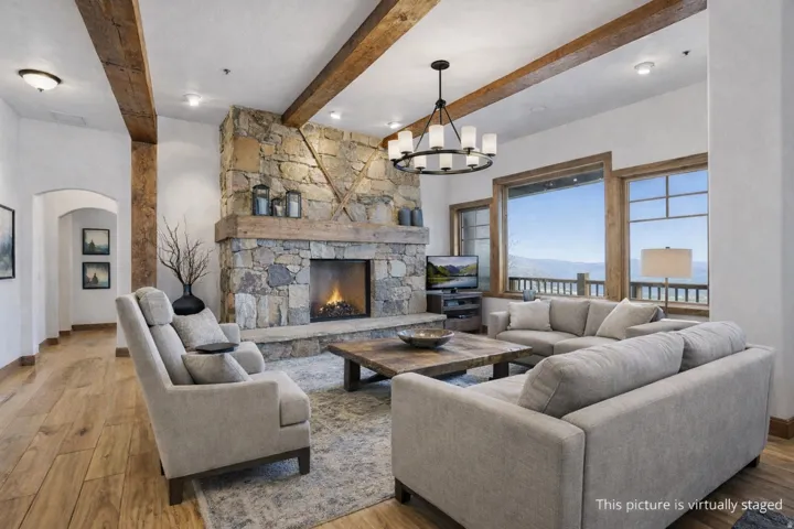 Spacious living area featuring a stone fireplace with a substantial wood mantel, exposed ceiling beams, wood-finish flooring, and expansive windows offering distant mountain scenery