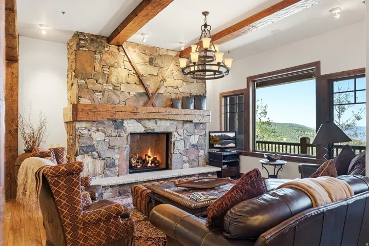 Living area featuring a stone fireplace with a thick wood mantel, exposed ceiling beams, wood-finish flooring, and expansive windows