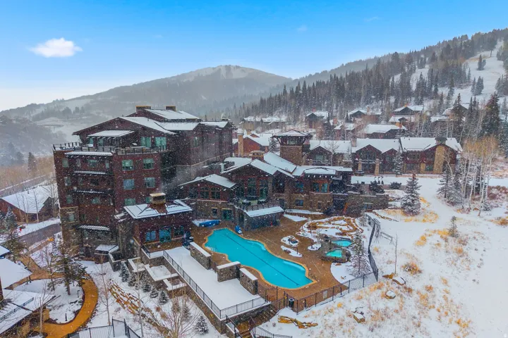 Architectural complex featuring a multi-story building with balconies, an outdoor pool, and two hot tubs, all surrounded by snow-covered grounds and evergreens