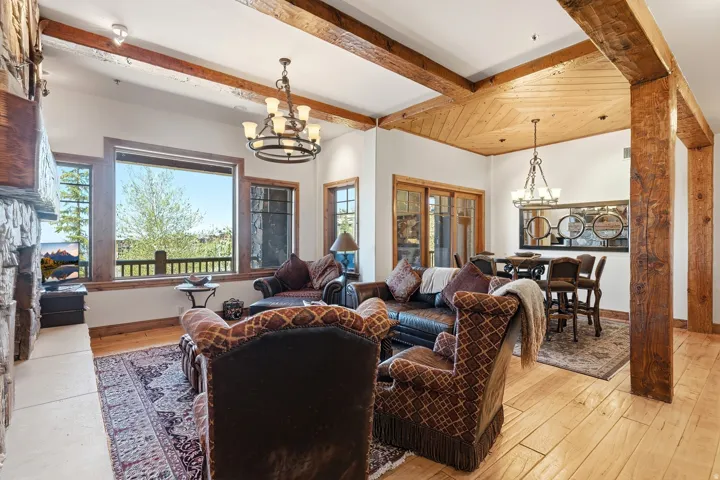 Open-concept living area featuring exposed wood beams, wood-finish flooring, a stone-clad fireplace, and large windows with integrated wood trim