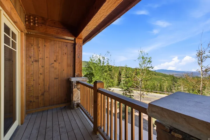 Wood-paneled outdoor balcony featuring timber beams, a stone-clad pillar, and a wood railing overlooking a tree-lined landscape with distant hills under a clear sky