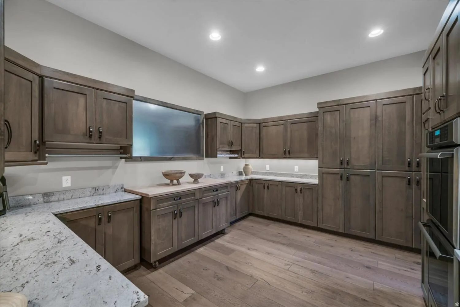 Kitchen with double oven, light wood-style flooring, dark brown cabinets, light stone countertops, and recessed lighting