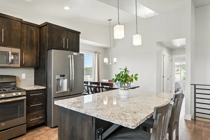 Kitchen featuring stainless steel appliances, pendant lighting, light stone countertops, dark brown cabinetry, and dark wood finished floors