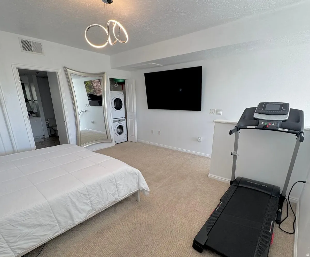 Bedroom featuring light carpet, a textured ceiling, estacked washer and dryer, a chandelier, and ensuite bath