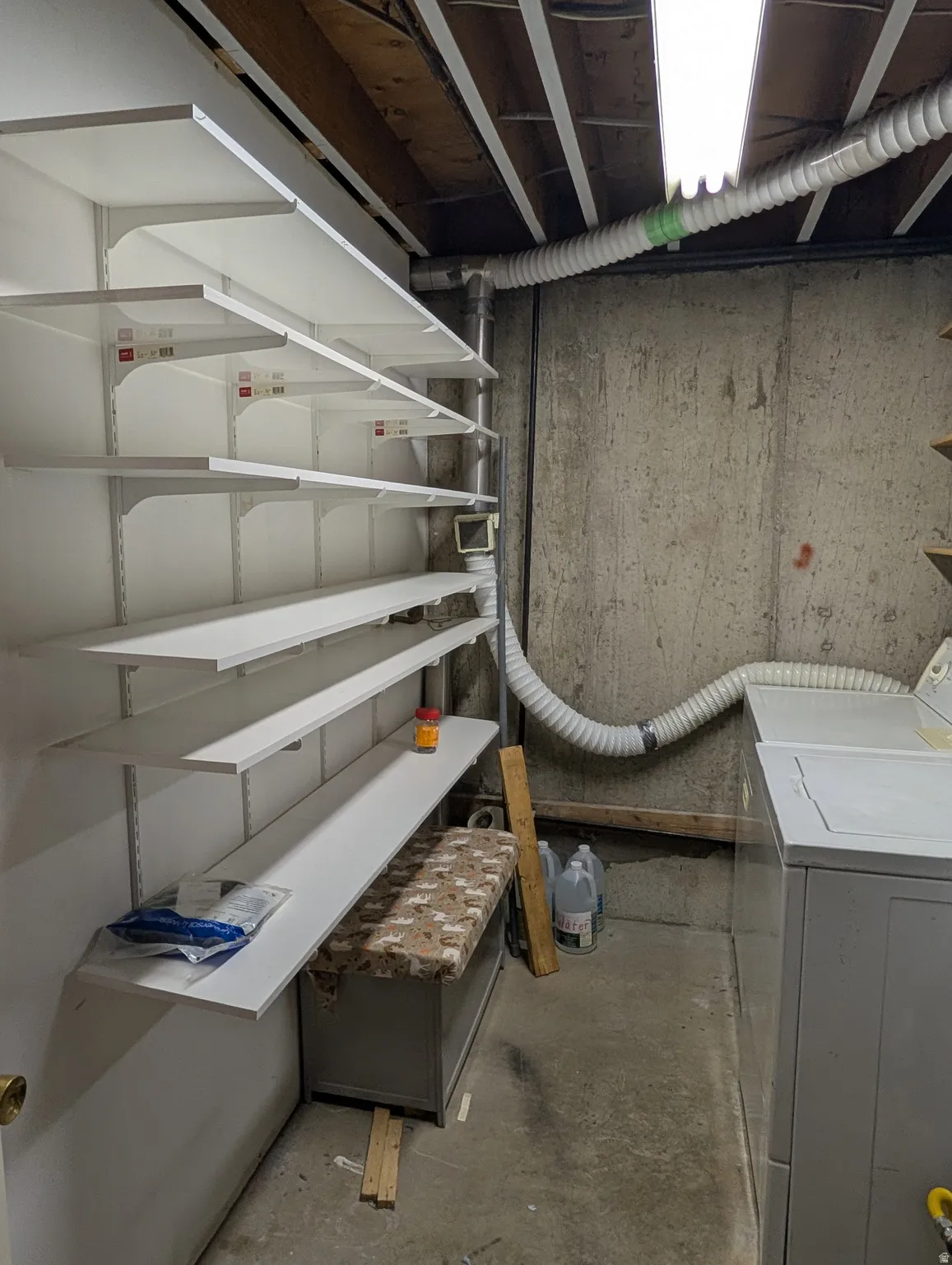 Utility area featuring concrete walls, exposed ceiling joists, white adjustable shelving, a fluorescent light fixture, and visible ductwork
