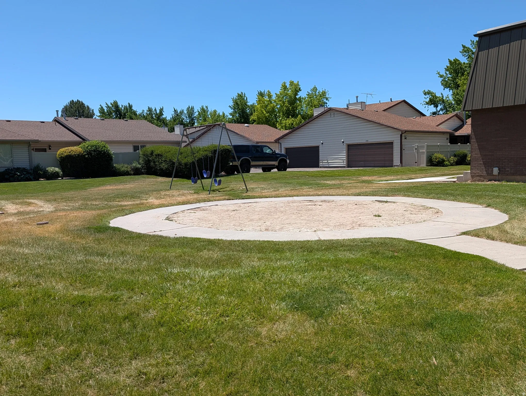 Community greenspace featuring a sand pit with a concrete border, a swing set, and a perimeter of mature trees