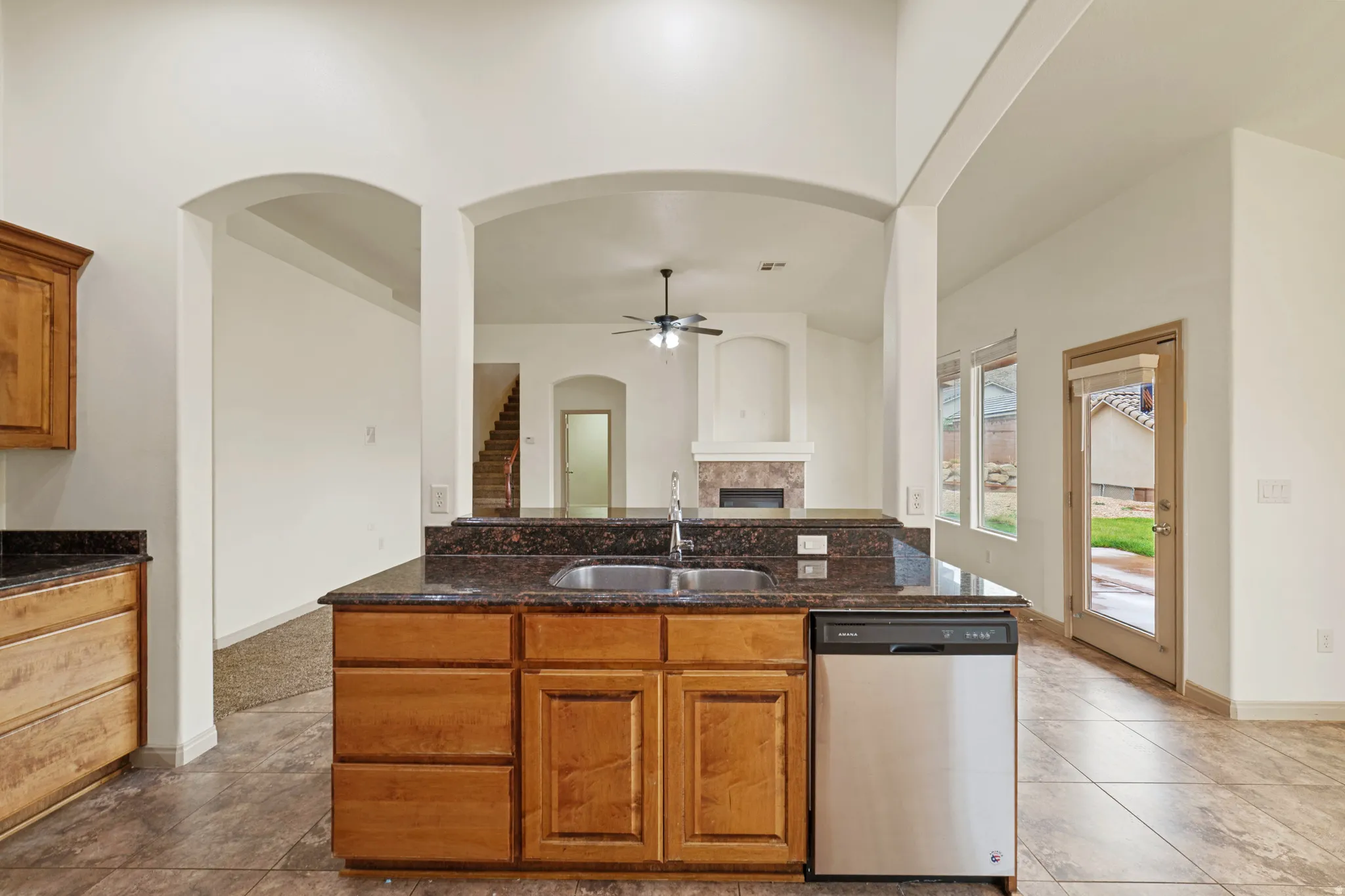 Kitchen with brown cabinetry, stainless steel dishwasher, ceiling fan, a tile fireplace, and dark stone counters