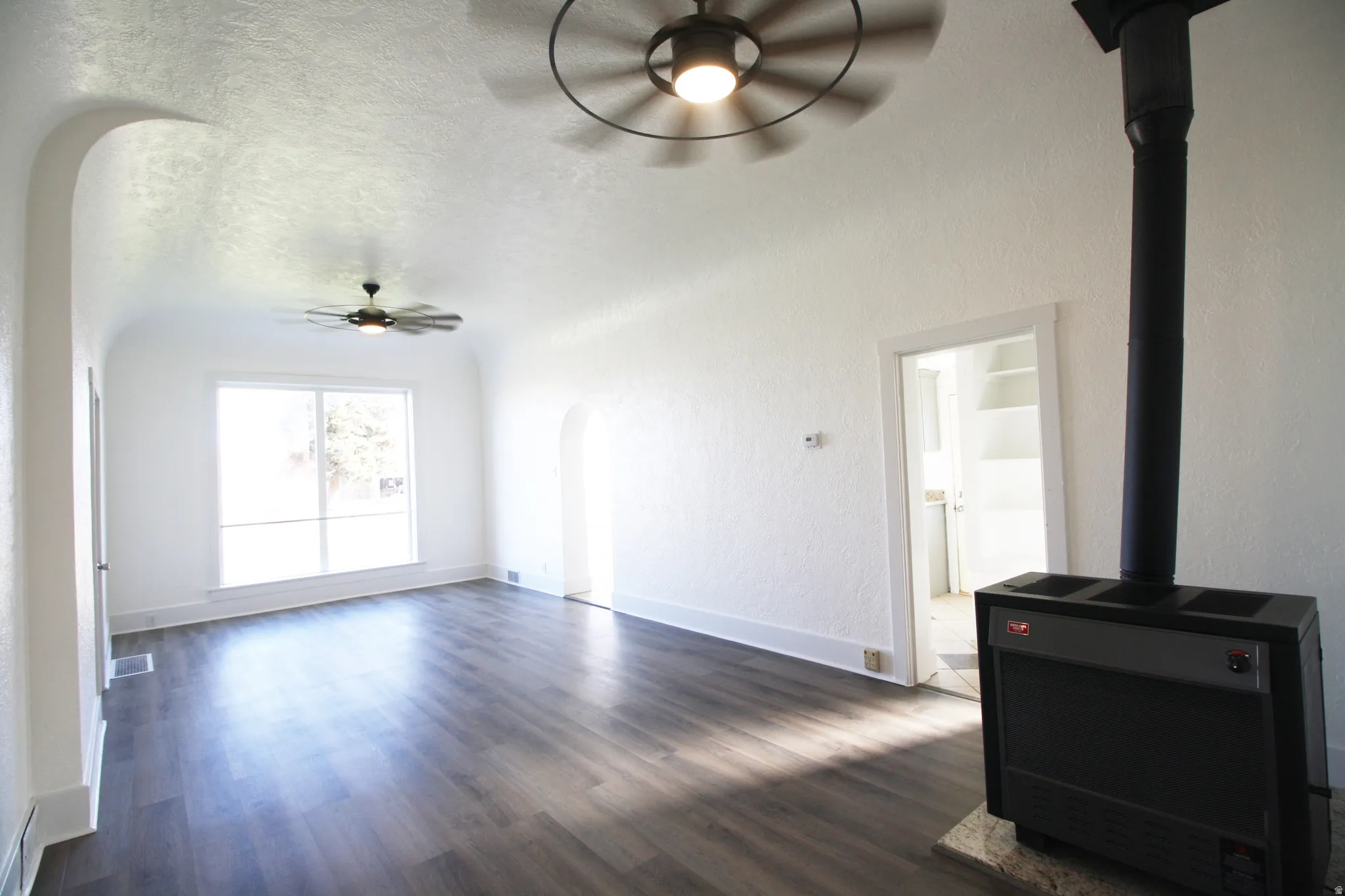 Family room featuring ceiling fan, a wood stove, arched walkways, dark wood-style floors, and a textured wall