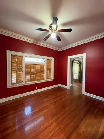 Interior space featuring wood-finish flooring, a ceiling fan with integrated lighting, crown molding, and a window with plantation shutters and an upper transom detail