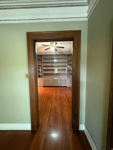 Archway with dark wood trim opening to a room featuring extensive built-in shelving, wood-finish flooring, a ceiling fan, and decorative crown molding