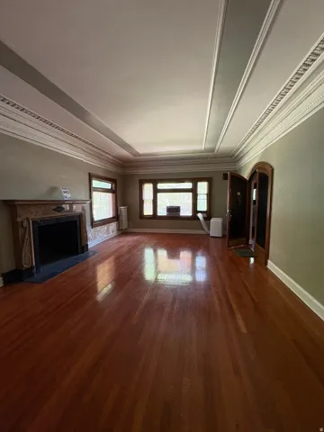 Spacious room featuring rich wood-finish flooring, an ornate fireplace with a stone mantel, detailed crown molding, a coffered ceiling, and a prominent arched entryway