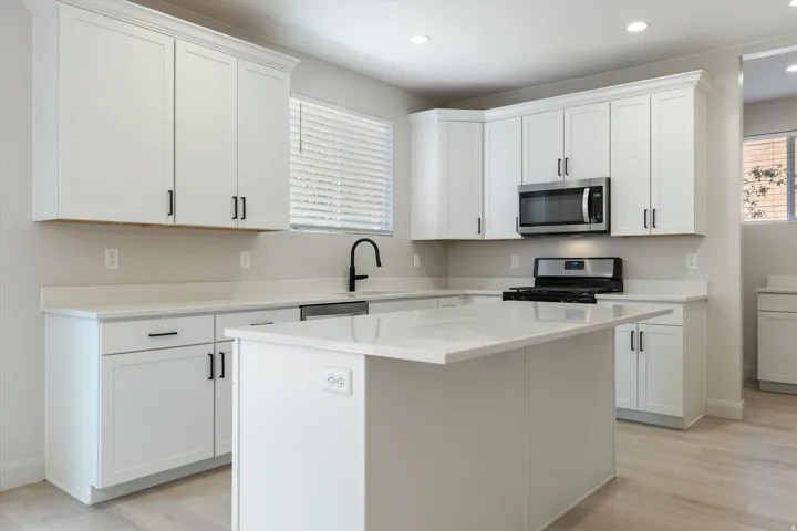 White shaker cabinetry with black hardware, stainless steel appliances, light-toned countertop surfaces, a central island, and wood-finish flooring