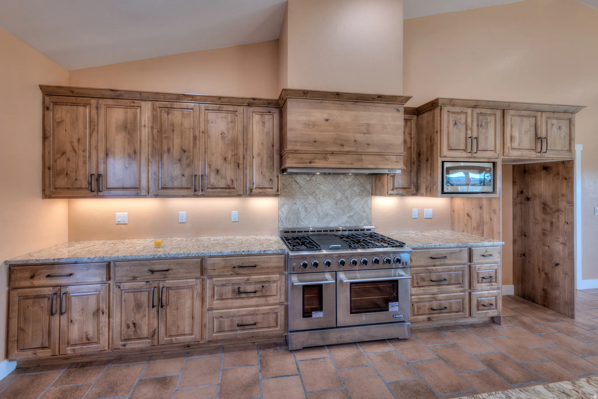 Kitchen with stainless steel appliances, lofted ceiling, light stone countertops, decorative backsplash, and brown cabinetry