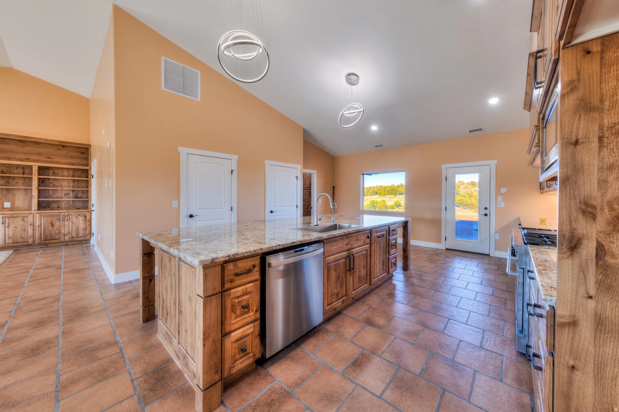 Kitchen featuring brown cabinets, decorative light fixtures, an island with sink, stainless steel appliances, and light stone countertops