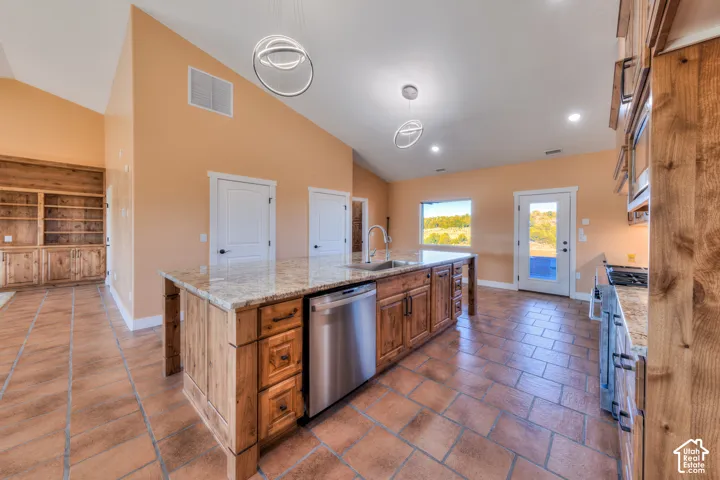 Kitchen featuring brown cabinets, decorative light fixtures, an island with sink, stainless steel appliances, and light stone countertops