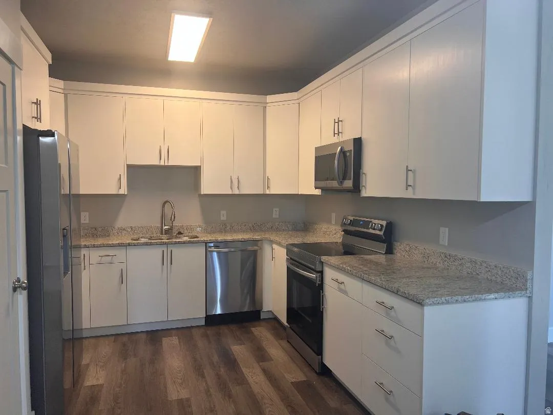 Kitchen with stainless steel appliances, light stone counters, white cabinets, and dark wood-style flooring