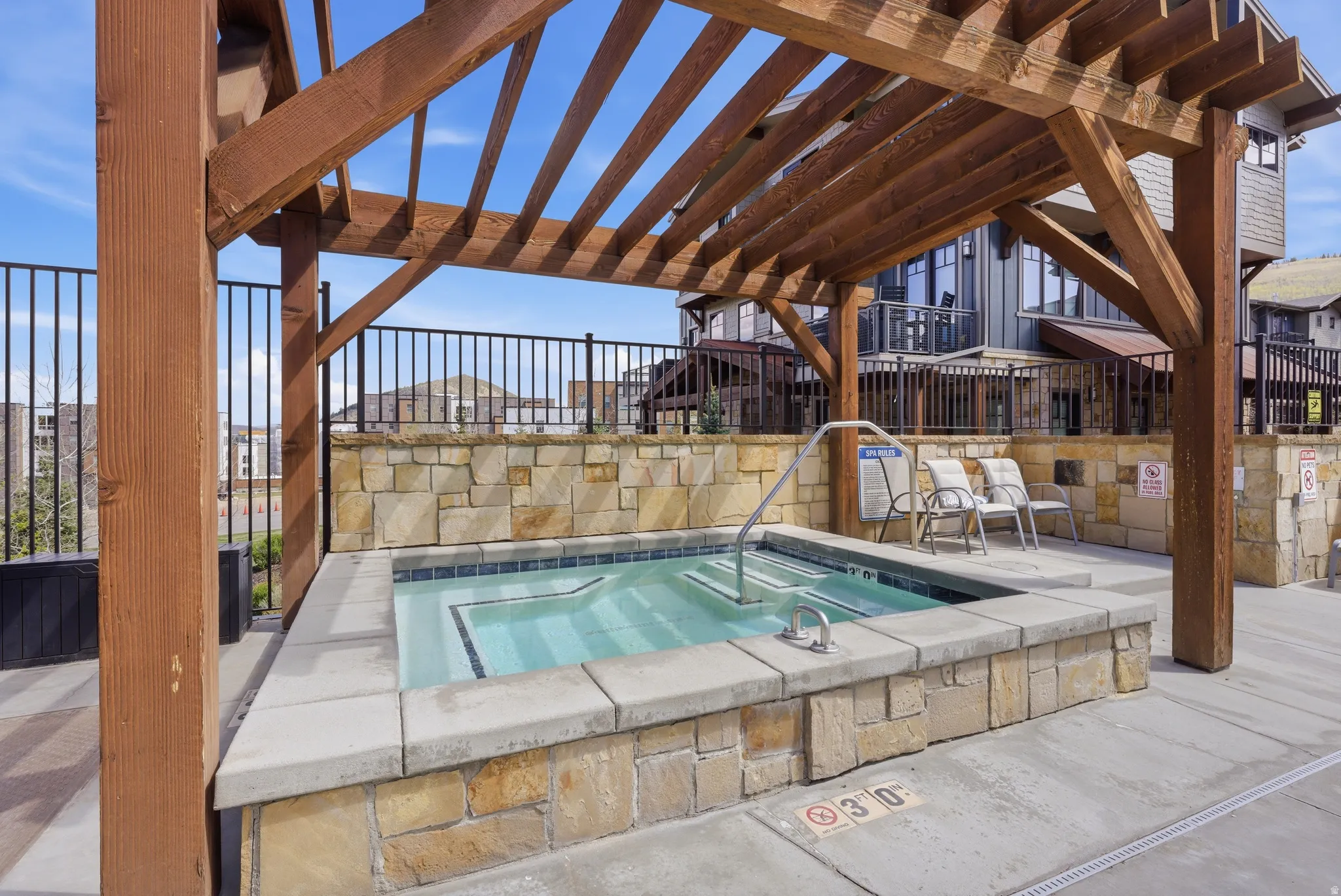 Outdoor spa with stone surround, concrete patio, wood pergola, and black metal fencing