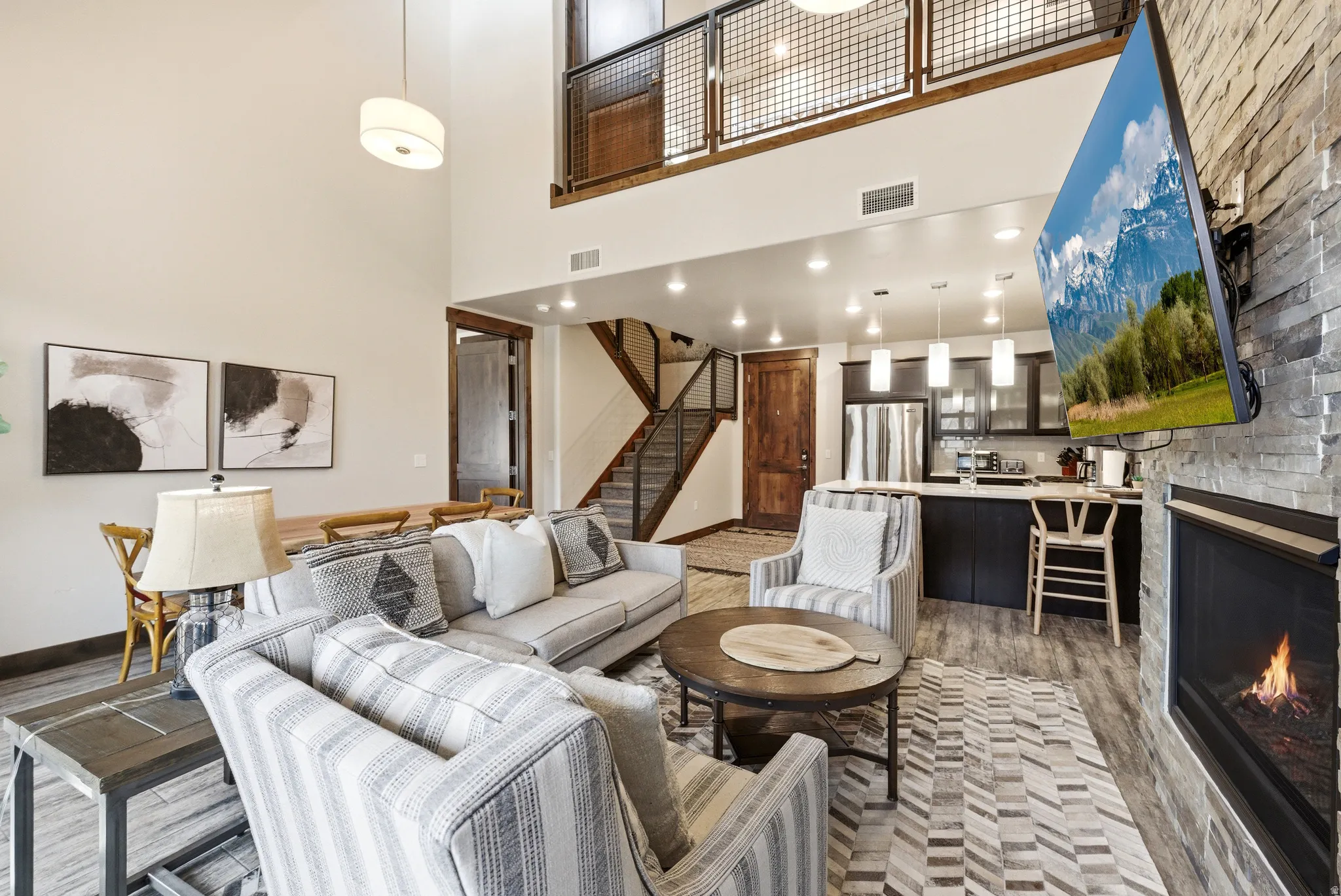Spacious open-concept living area featuring high ceilings, a stacked stone fireplace, wood-finish flooring, and a second-story interior balcony with metal railing