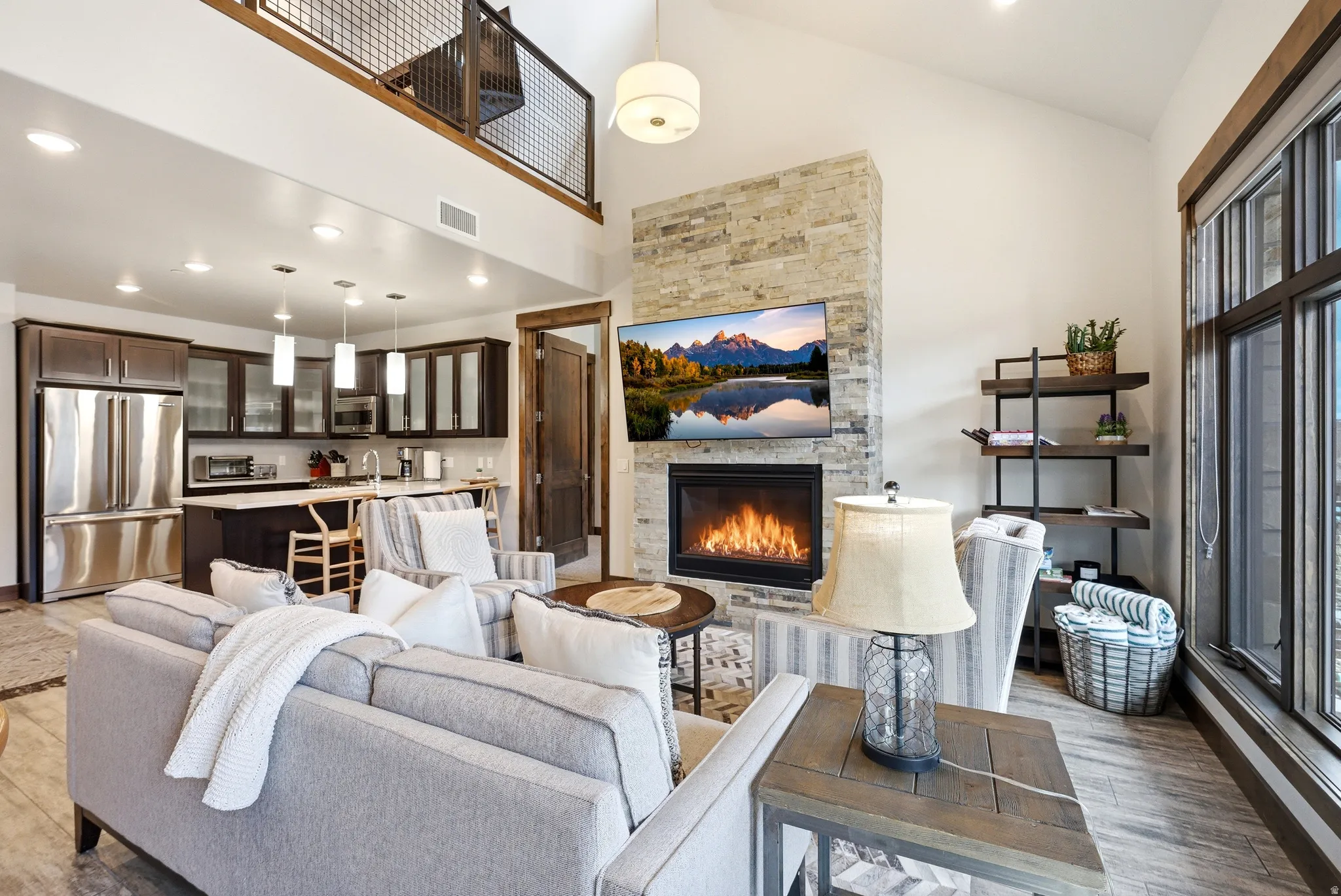 Vaulted ceiling living area featuring a stone-clad fireplace, large windows, wood-finish flooring, and a stainless steel appliance kitchen with pendant lighting