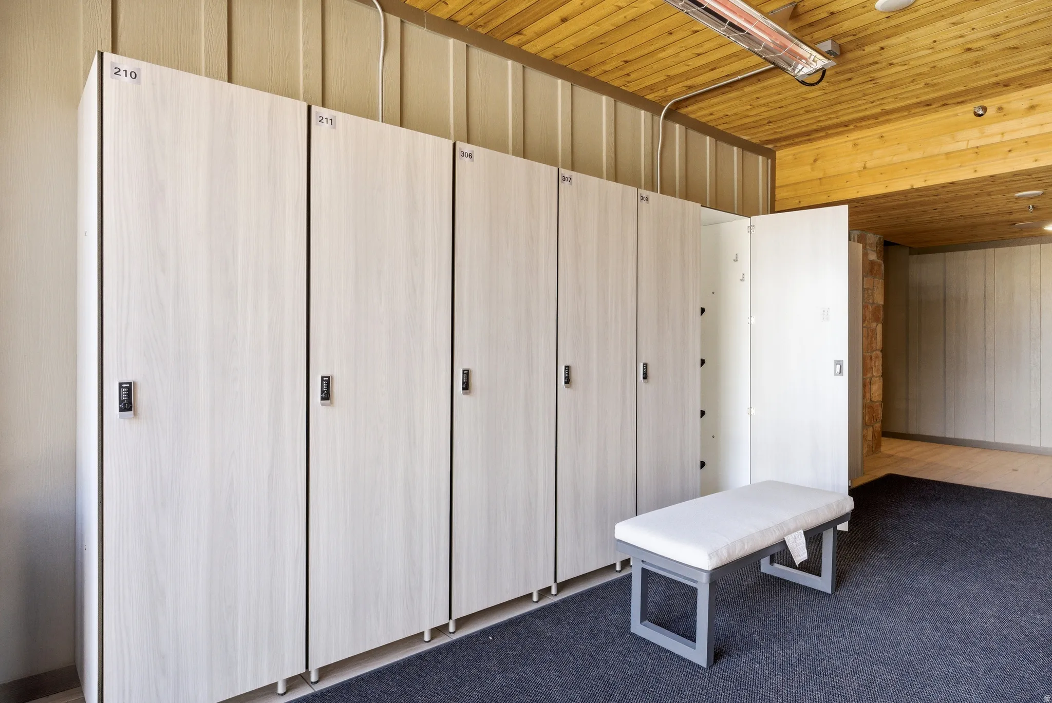 Locker room featuring multiple light wood-finish lockers with individual locks, a padded bench, and a textured dark gray floor covering