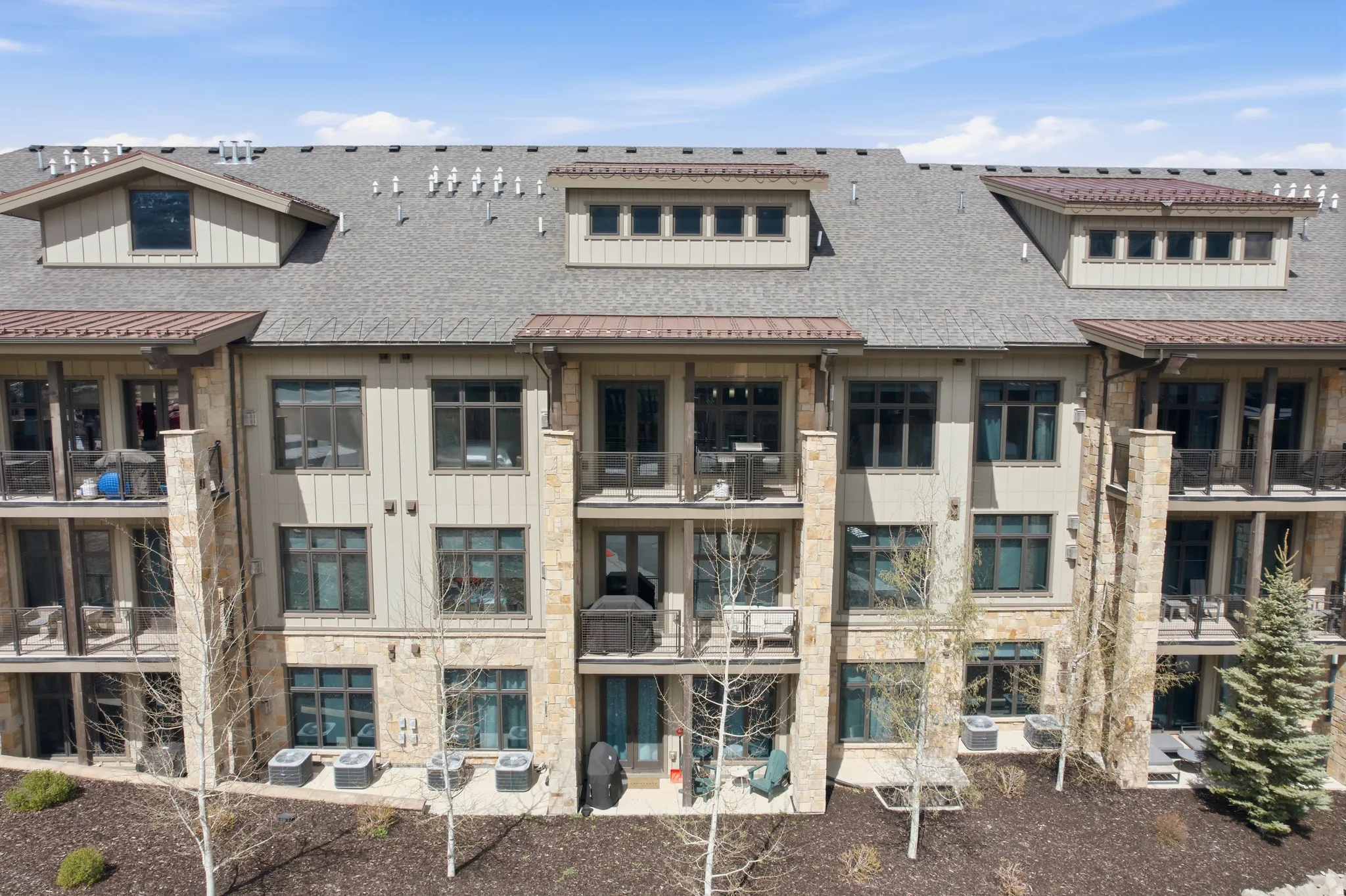 Multi-story condominium building featuring stone and siding exterior, multiple balconies, individual patios, and dormer windows