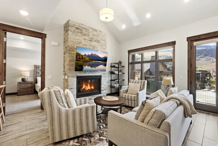 Living area featuring a stone-clad fireplace with a mounted television, a vaulted ceiling, wood-finish flooring, a wall of windows, and a glass-paned door with dark wood trim