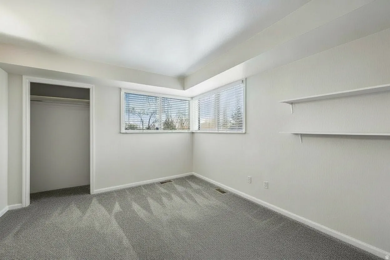 Bright room featuring an expansive window with blinds, soft gray patterned carpeting, recessed closet space, and two built-in wall shelves