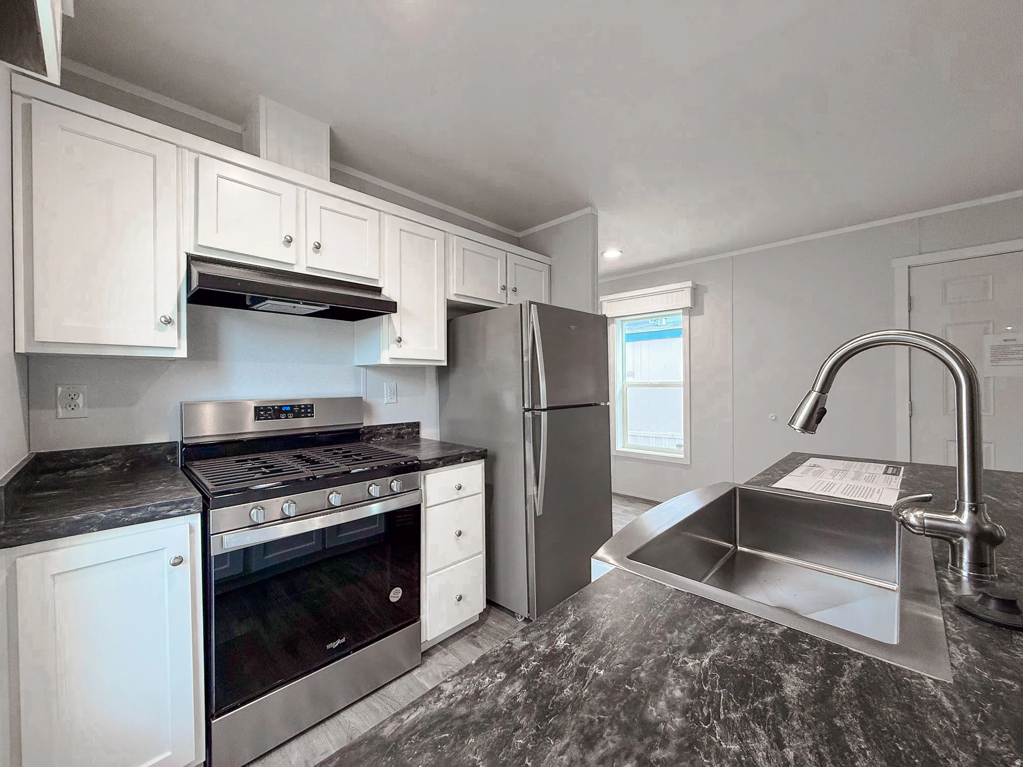 Kitchen with stainless steel appliances, under cabinet range hood, white cabinetry, ornamental molding, and dark stone counters