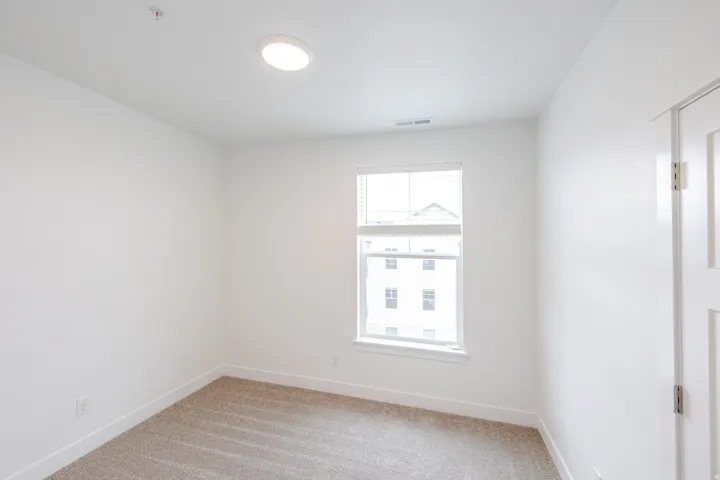 Bright room featuring a single window with blinds, neutral carpeting, white walls, and a flush-mount ceiling light