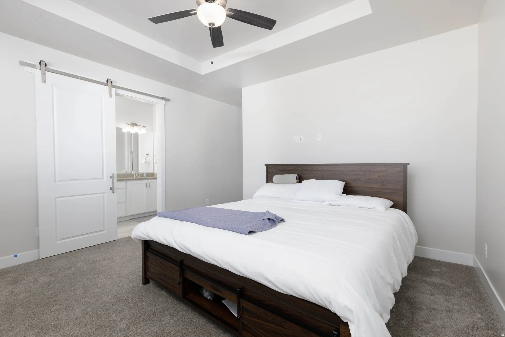 Carpeted bedroom with a raised ceiling, a barn door, connected bathroom, and ceiling fan