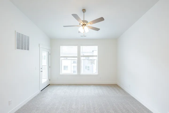 Spacious room featuring light gray carpet flooring, two large windows with blinds, a ceiling fan with integrated lighting, and a glass-paneled door opening to a balcony with a black metal railing