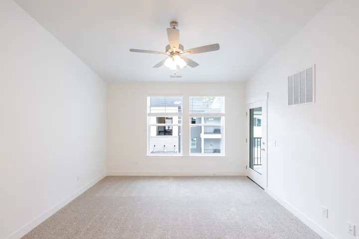 Spacious room featuring neutral carpeting, white walls, a ceiling fan with light fixture, large windows, and a glass-paneled door leading to an exterior balcony