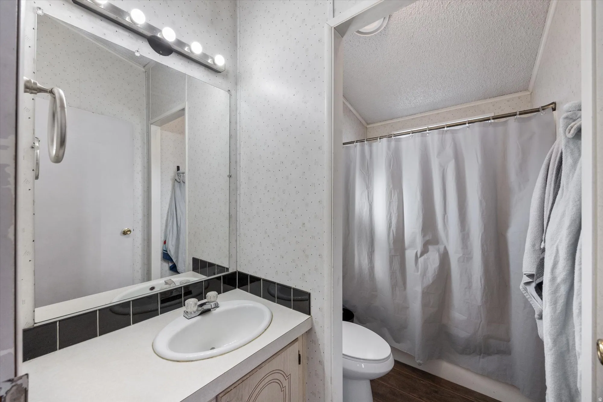 Full bath featuring a textured ceiling, vanity, wallpapered walls, dark wood finished floors, and shower / tub combo with curtain