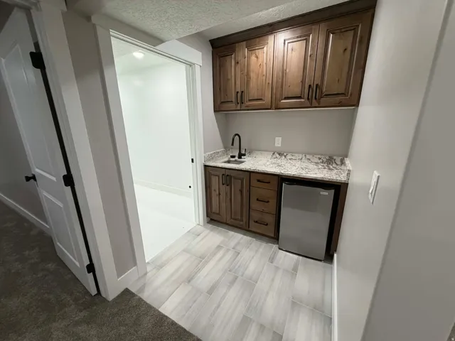Indoor wet bar with a textured ceiling, light stone counters, stainless steel refrigerator, and dark brown cabinets