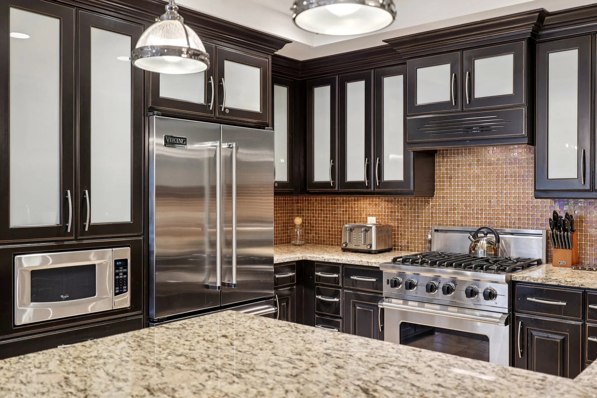 Kitchen featuring premium appliances, tasteful backsplash, glass insert cabinets, light stone counters, and dark brown cabinets