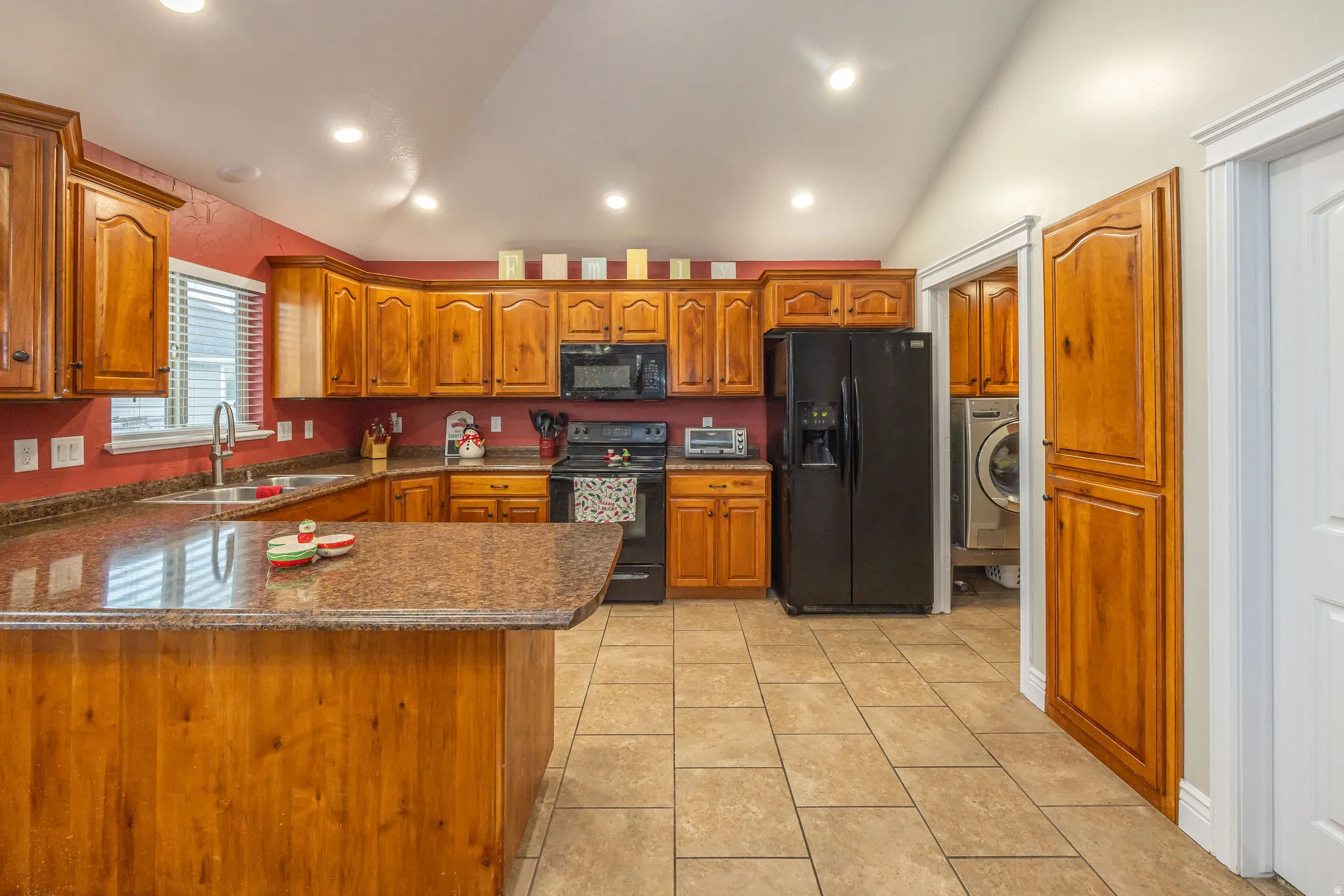 Kitchen featuring black appliances, brown cabinetry, light tile patterned flooring, vaulted ceiling, and a peninsula