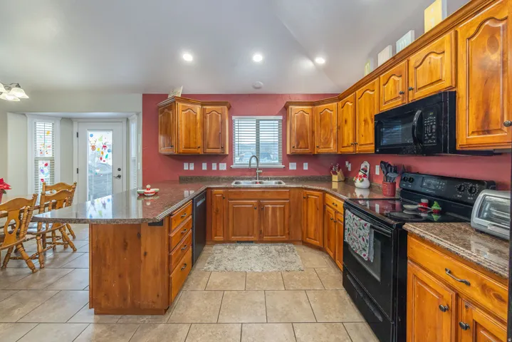 Kitchen featuring dark stone counters, black appliances, brown cabinets, a peninsula, and vaulted ceiling