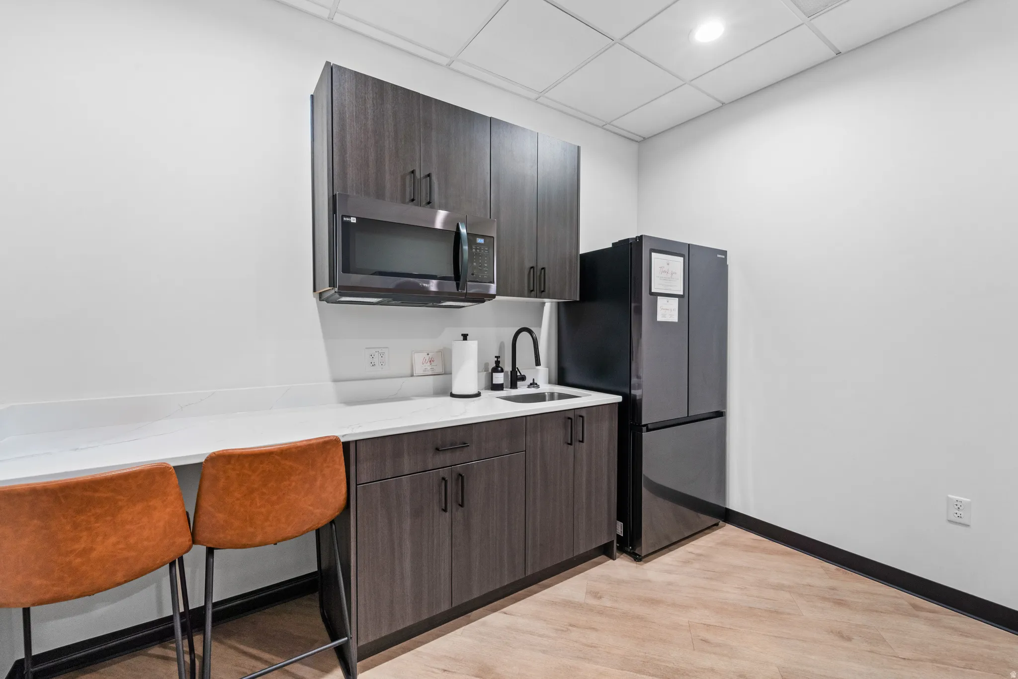 Kitchen with a kitchen bar, stainless steel microwave, freestanding refrigerator, dark brown cabinetry, and a drop ceiling