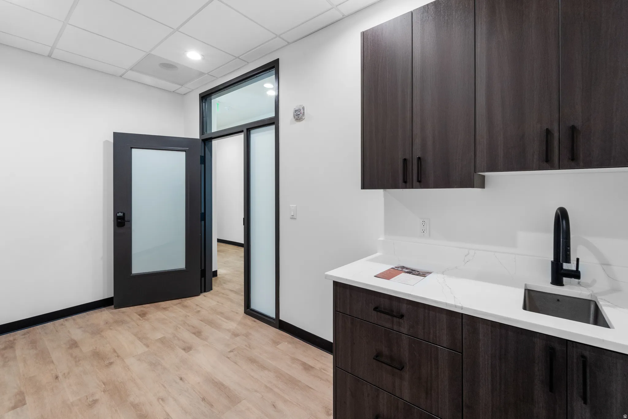 Kitchen with dark brown cabinetry, modern cabinets, a drop ceiling, light wood-style floors, and light stone counters